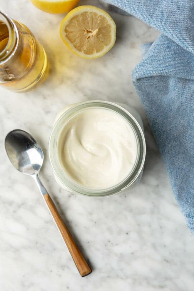 Einfaches Selbstgemachtes Mayonnaise Rezept 2 Ein Blick von oben auf goldgelbe, cremige Mayonnaise in einem Glas, daneben ein Löffel, frische Zitronenhälften, eine Flasche Avocadoöl und ein hellblaues Leinentuch auf dem Tisch.