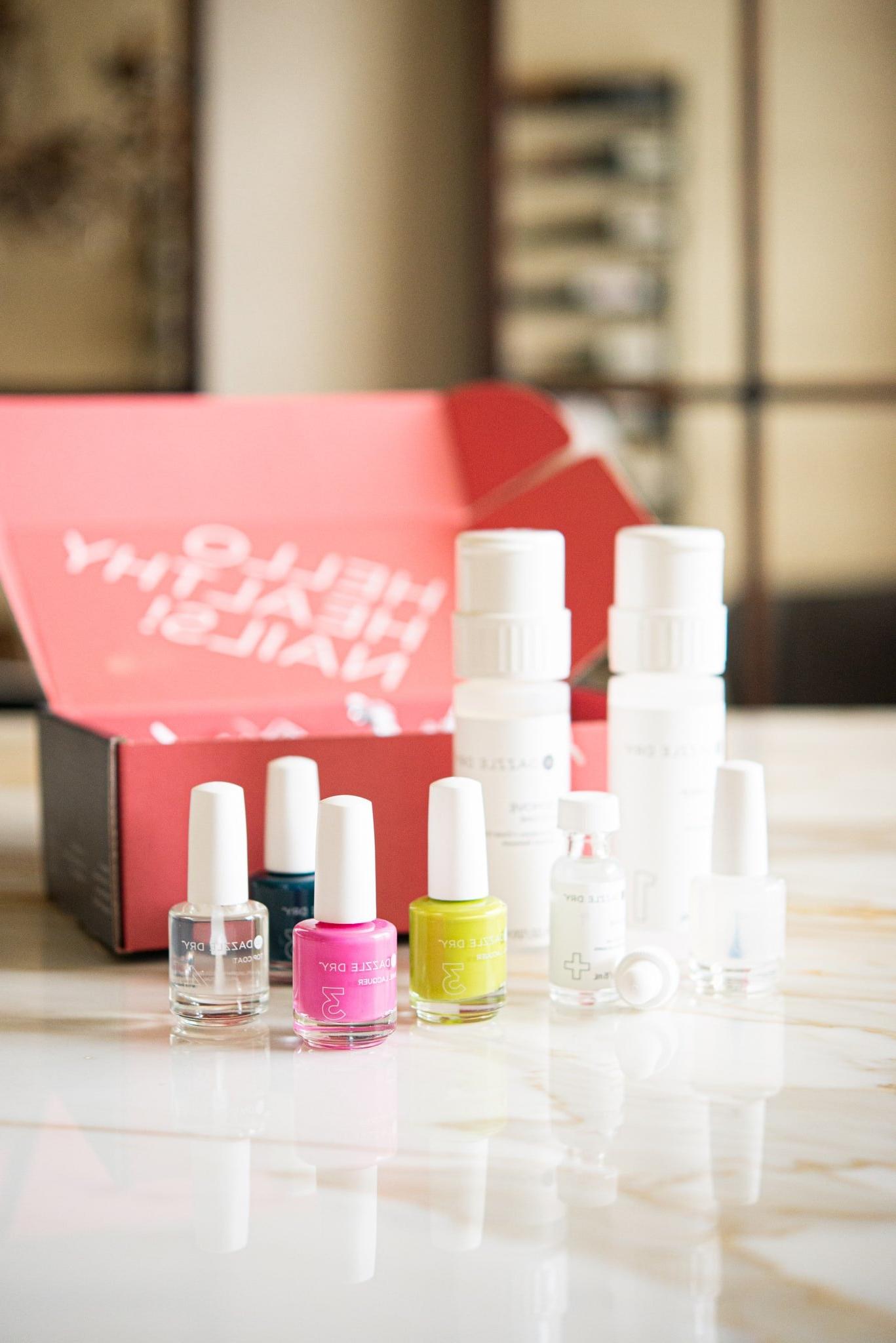 Dazzle Dry Nagelset mit drei Nagellacken, einem Überlack und einem Entferner, positioniert vor einer leuchtend rosafarbenen Box – ein Blickfang für jede Nagelpflege-Sammlung.