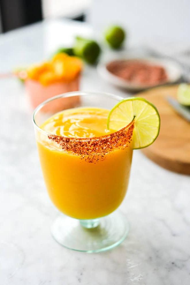 Einfaches Mango-Margarita-Rezept (Gefroren und auf Eis) 2 Ein Glas Frozen Mango Margarita mit Chilischnee-Rand, Zutaten für Mango Margaritas im Hintergrund
