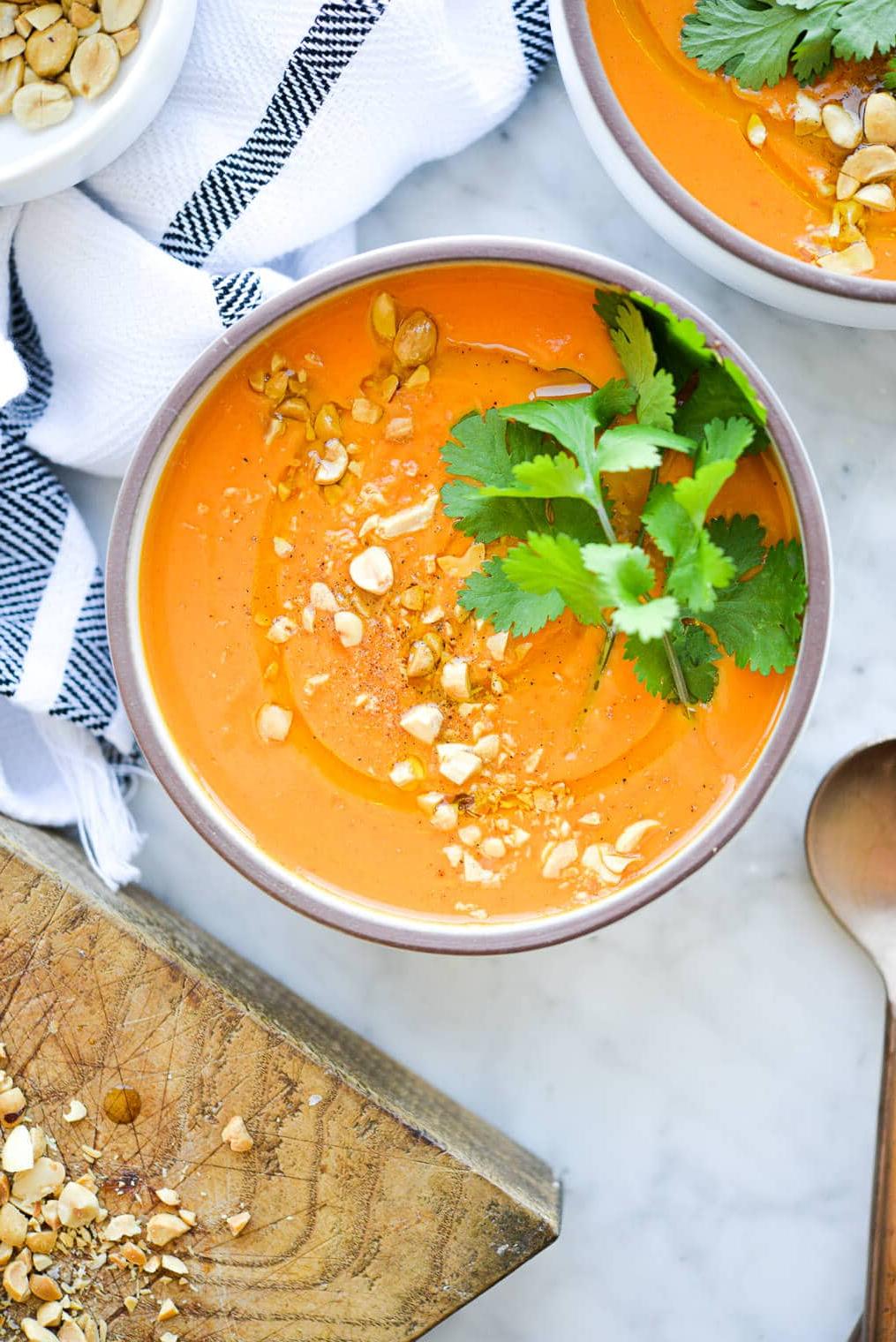 Thailändische Karotten-Ingwer-Suppe 2 Eine Schüssel mit leuchtend oranger Thai-Karottensuppe, garniert mit frischem Koriander und zerdrückten Erdnüssen, auf einer Marmoroberfläche.
