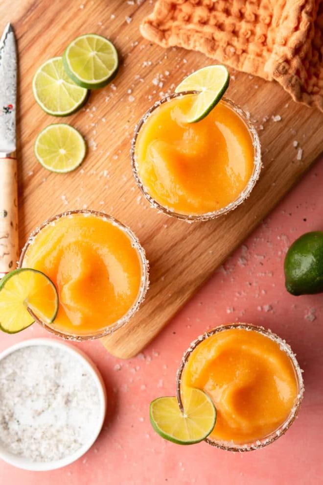 Das beste Pfirsich-Margarita-Rezept 2 Aufnahme von oben: Gefrorene Pfirsich Margaritas in Gläsern, garniert mit Meersalzflocken und einer Limettenscheibe, auf einer pinkfarbenen Oberfläche mit Schneidebrett, Schälmesser, einer Prise Meersalzflocken, Limettenscheiben und orangefarbenem Leinentuch.