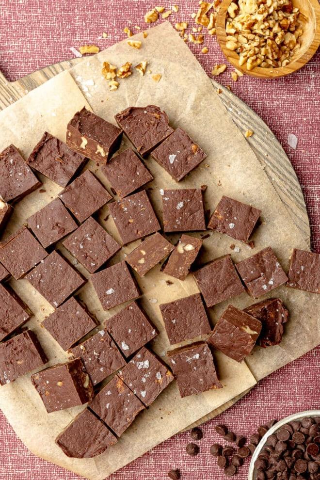 Schokoladen-Walnuss-Fudge in kleinen Quadraten auf braunem Backpapier auf einer dunkelrosafarbenen Tischdecke, daneben eine Holzschale mit Walnüssen und eine kleine Schüssel mit Schokoladenstückchen.