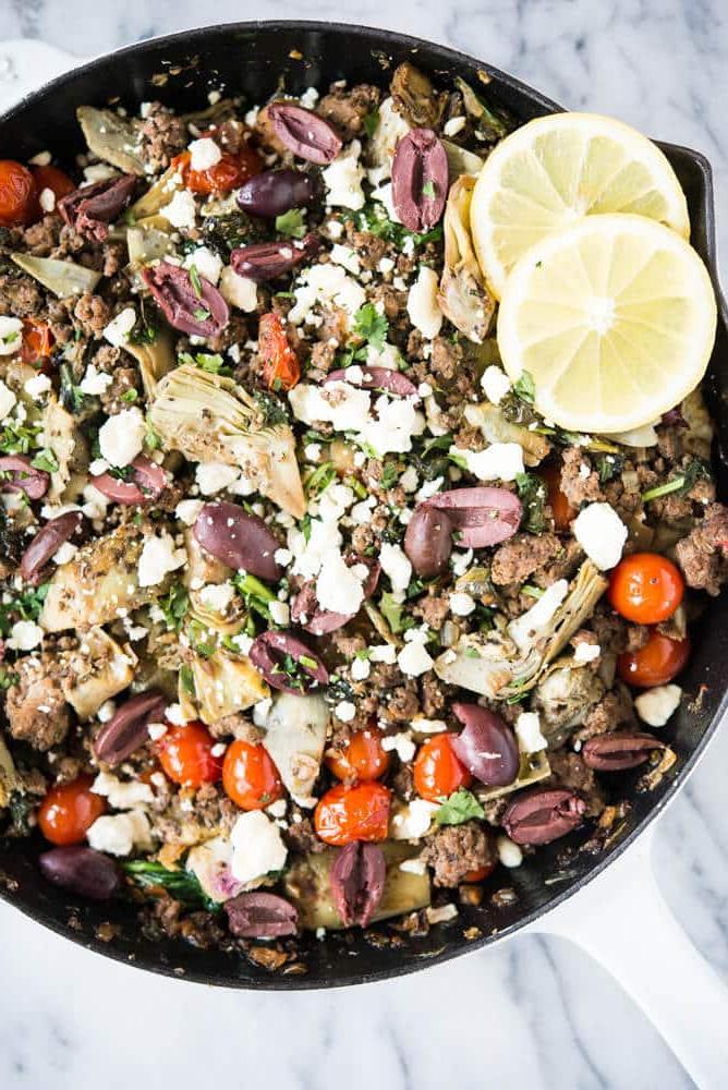Mediterrane Pfanne mit Spinat, Feta, Oliven und Artischocken in einer Gusseisenpfanne auf einer Marmoroberfläche