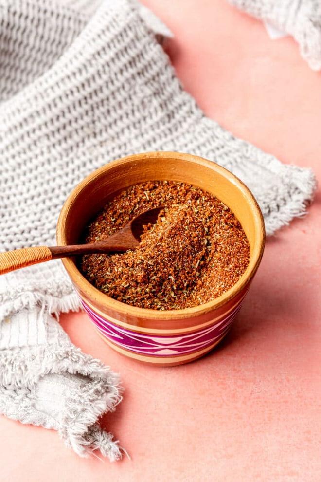 Beste Trockenmarinade für Schweinefilet 2 Eine braune Tonschale mit fuchsiafarbenen und weißen Mustern, gefüllt mit einer Gewürzmischung, daneben ein kleiner Holzlöffel. Die Schale steht auf einer rosa Oberfläche mit einem hellgrauen, strukturierten Leinen.