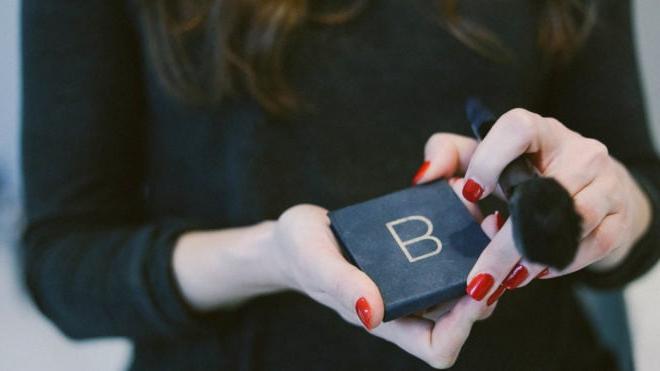 Eine Frau mit dunklem, langem Haar und roten Fingernägeln, die eine Rouge-Palette und einen Rougepinsel für ihre minimalistische Make-up-Routine hält.
