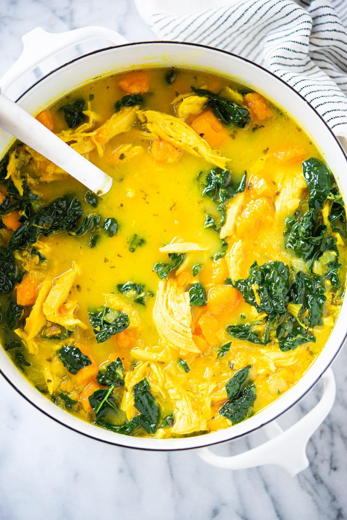 Hühnersuppe mit Ingwer und Kurkuma 2 Gelbe Kurkuma-Hühnersuppe mit Süßkartoffeln und Grünkohl in einem weißen Topf auf einer Marmoroberfläche