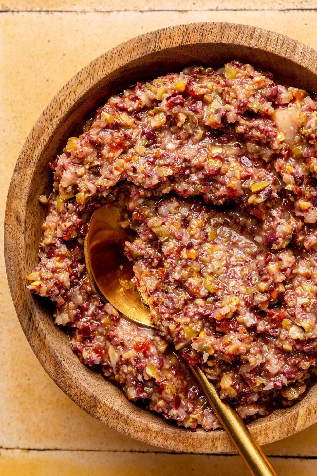 Oliven-Tapenade 2 Oliven-Tapenade in einer hölzernen Servierschale. Ein goldfarbener Löffel liegt daneben.