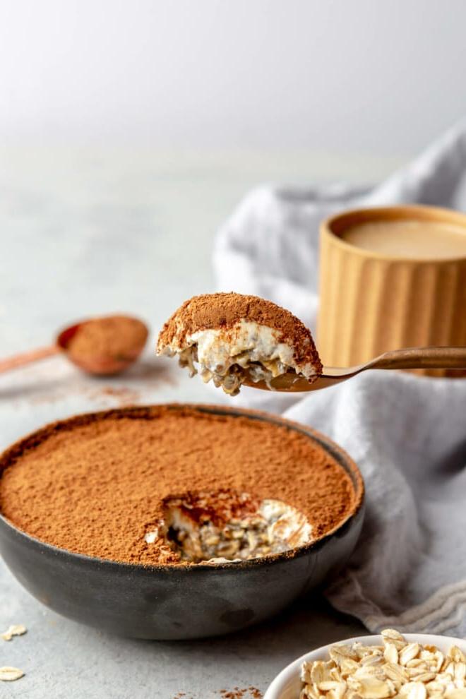 Tiramisu Overnight Oats 2 Eine seitliche Ansicht einer Schale Tiramisu Overnight Oats in einer schwarzen Schale. Eine Hand hält den Löffel mit einem Löffel Haferflocken über der Schale. Im Hintergrund ist eine Tasse Kaffee zu sehen.