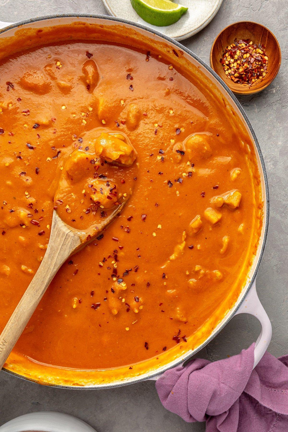 Ein weißer Topf mit Kürbis-Curry, das mit roten Pfefferflocken bestreut ist. Ein Holzlöffel ist zu sehen, wie er einen Teil des Currys schöpft.