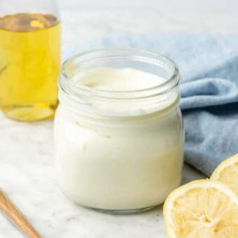 Einfaches Selbstgemachtes Mayonnaise Rezept 5 Glas mit hausgemachter Mayonnaise auf einer grau-weißen Theke mit Zitronenhälften, Avocadoöl im Hintergrund, einem Servierlöffel und einem hellblauen Leinentuch.