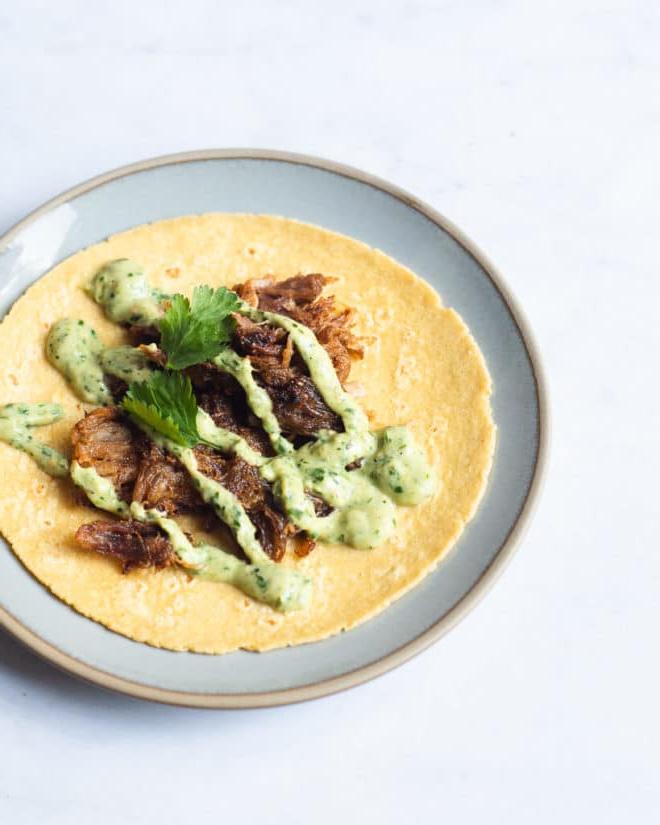Ein einzelner Carnitas Taco, belegt mit cremiger Avocado-Verde-Sauce und Koriander, offen auf einem kleinen blauen Teller