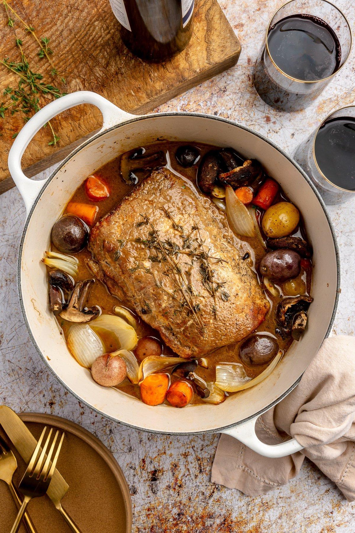 **Balsamico-Schweinelendenrezept** 11 Ein fertig gegarter Balsamico-Schweinebraten liegt in einem weißen, emaillierten Topf. Zwei Weingläser, ein brauner Teller mit goldenem Besteck und ein Schneidebrett stehen daneben.