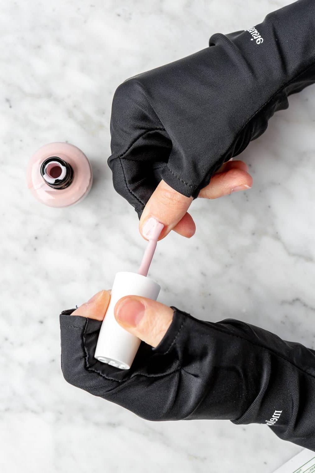220831_Gel Manicure-22 Hände tragen schwarze, fingerlose Handschuhe und tragen eine Schicht hellrosa Nagellack auf die Nägel auf.