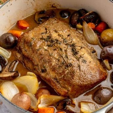 **Balsamico-Schweinelendenrezept** 12 Ein fertig gegarter Balsamico-Schweinebraten liegt in einem weißen, emaillierten Topf. Zwei Weingläser, ein brauner Teller mit goldenem Besteck und ein Schneidebrett stehen daneben.