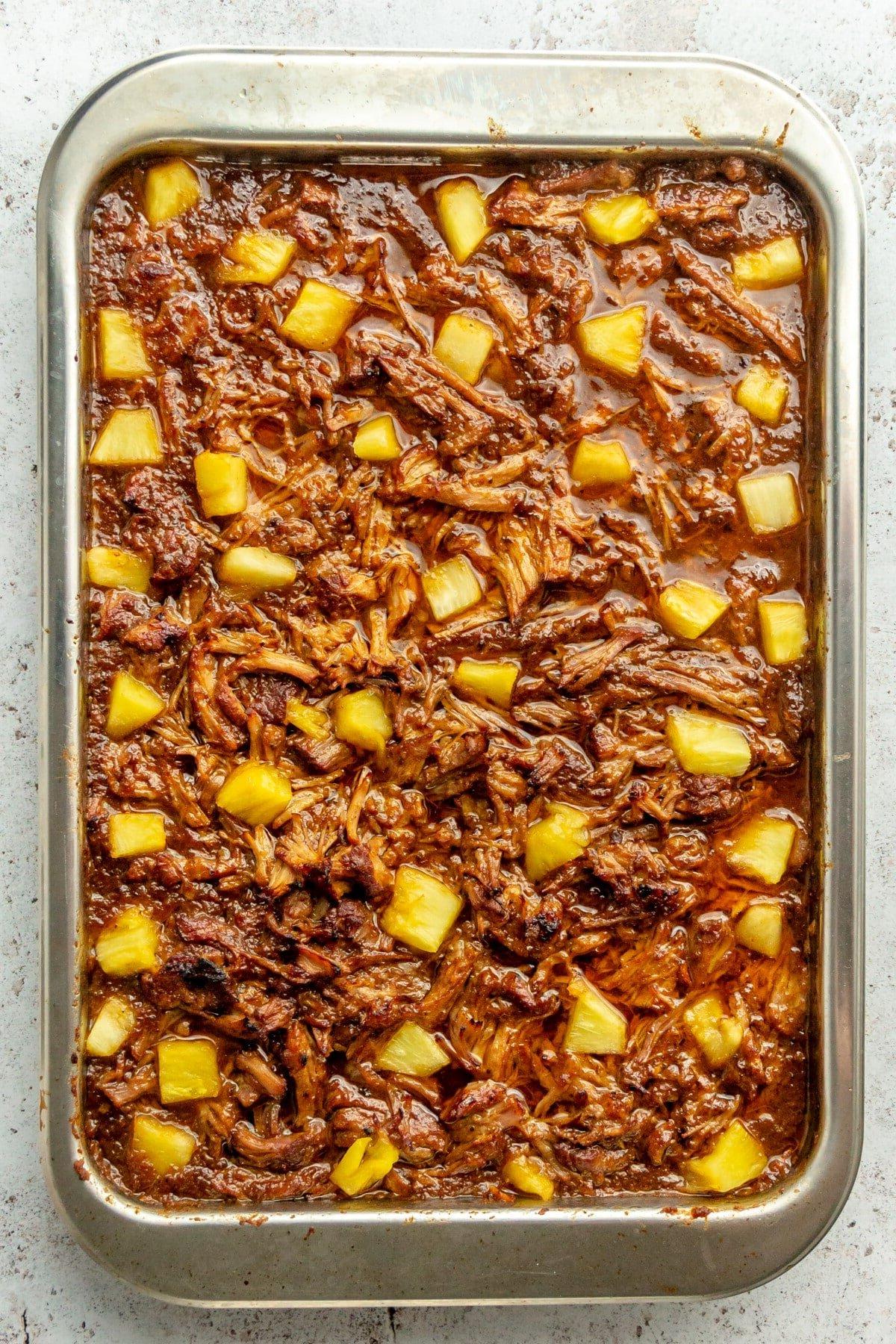 Tacos Al Pastor Rezept 13 Gegrilltes, geschreddertes mariniertes Schweineschulterfleisch und Ananasstücke auf einem Backblech auf hellem Hintergrund.