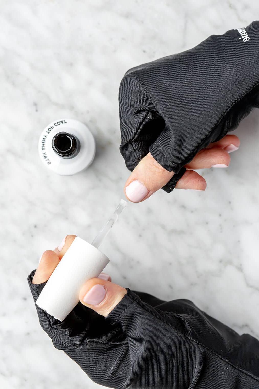 220831_Gel Manicure-17 Hände tragen fingerlose schwarze Handschuhe und tragen eine Deckschicht Nagellack auf die lackierten Nägel auf.