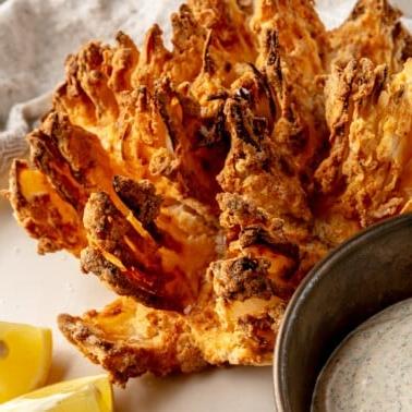 Heißluftfritteuse Blühende Zwiebel 13 Eine blühende Zwiebel aus der Heißluftfritteuse liegt perfekt gegart und goldbraun auf einem Teller, daneben eine Schale mit cremiger Dip-Soße und frischen Zitronenspalten für eine appetitliche Präsentation.