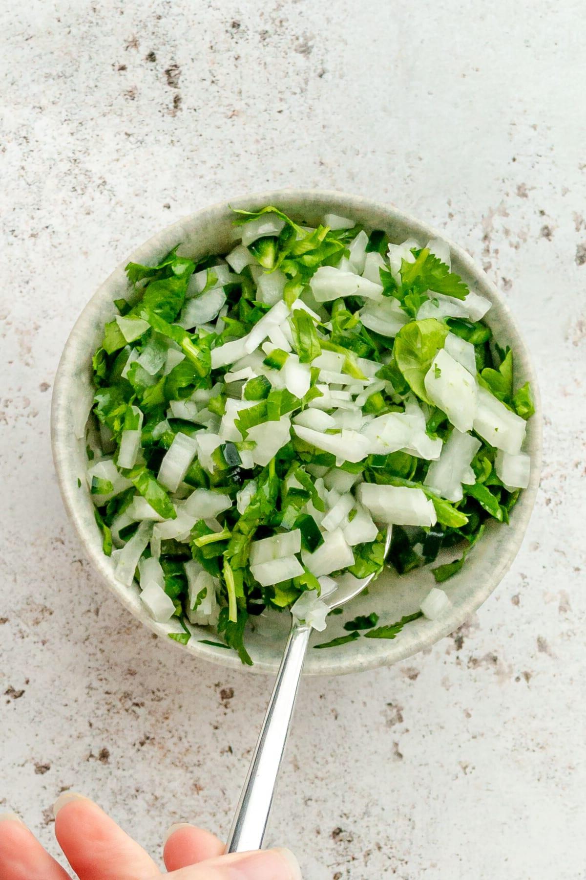 Tacos Al Pastor Rezept 14 Eine Zwiebel-Koriander-Jalapeño-Salsa wird in einer kleinen blauen Keramikschüssel auf hellem Hintergrund umgerührt.