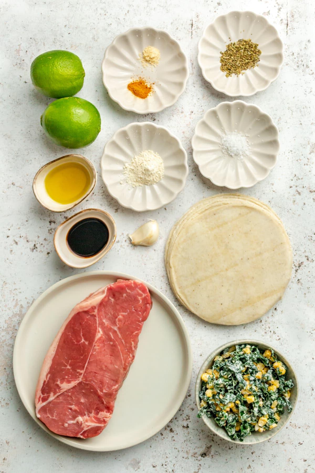 Carne Asada Tacos Rezept mit Adobo-Limetten-Marinade 3 Zutaten für Carne Asada Tacos mit Adobo-Limetten-Marinade liegen auf verschiedenen kleinen Tellern und Schalen neben einem Grünkohl-Mais-Krautsalat auf einer hellgrauen Oberfläche.