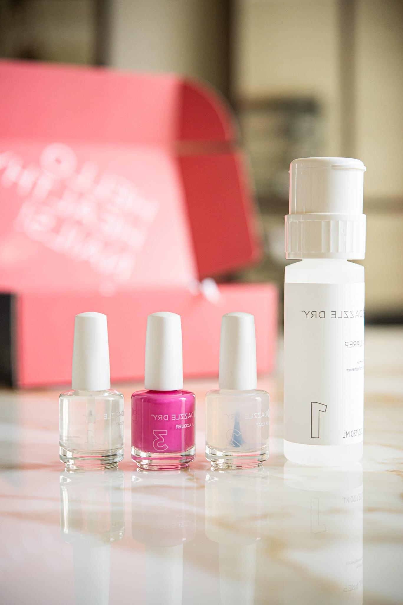Dazzle Dry Nail System Rezension 3 Dazzle Dry Nagelsystem – ein Arrangement aus leuchtend pinkem Nagellack neben zwei klaren Nagellacken und einem passenden Entferner, kunstvoll platziert auf einem eleganten Marmortisch.
