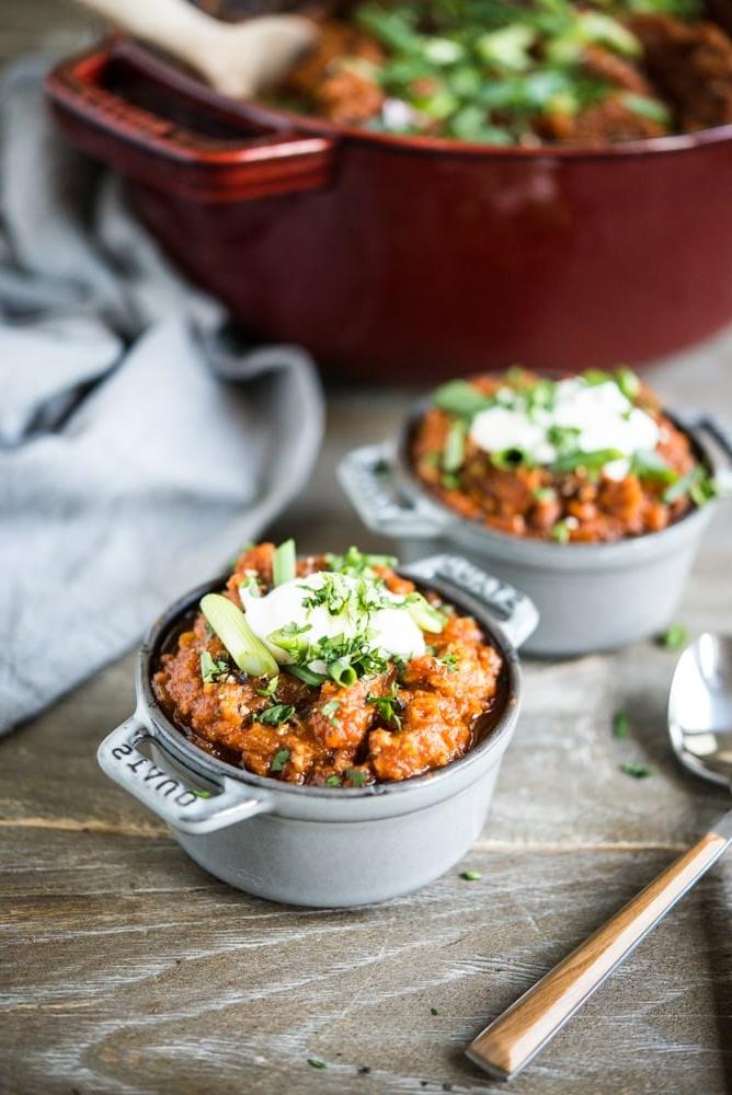Ein großer roter Emailletopf gefüllt mit einem tief orangefarbenen Kürbis-Chili, garniert mit geschnittenen Frühlingszwiebeln, daneben zwei individuelle Portionen Chili, garniert mit einem Klecks Sauerrahm und geschnittenen Frühlingszwiebeln.