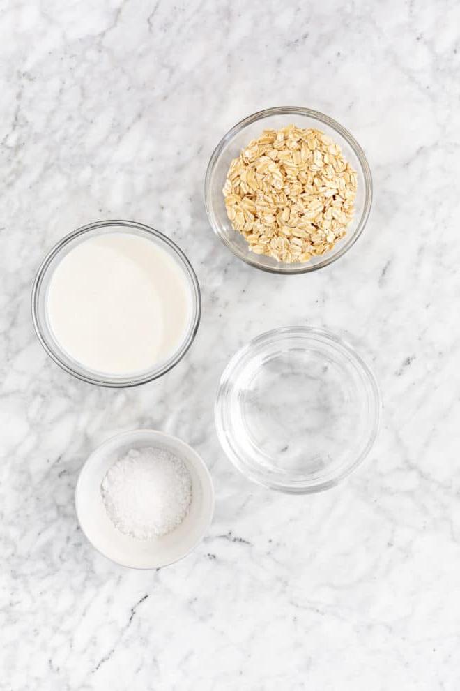Wie man Haferbrei auf dem Herd zubereitet 3 Stovetop oatmeal ingredients in glass bowls on a gray and white marble surface.