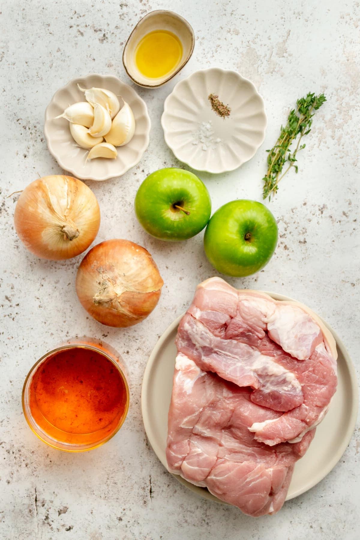 Apfelwein-Schweinebraten 3 Zutaten für Schweinefiletbraten mit Apfelwein, in verschiedenen Schüsseln und auf Tellern auf einer hellgrauen Oberfläche arrangiert.