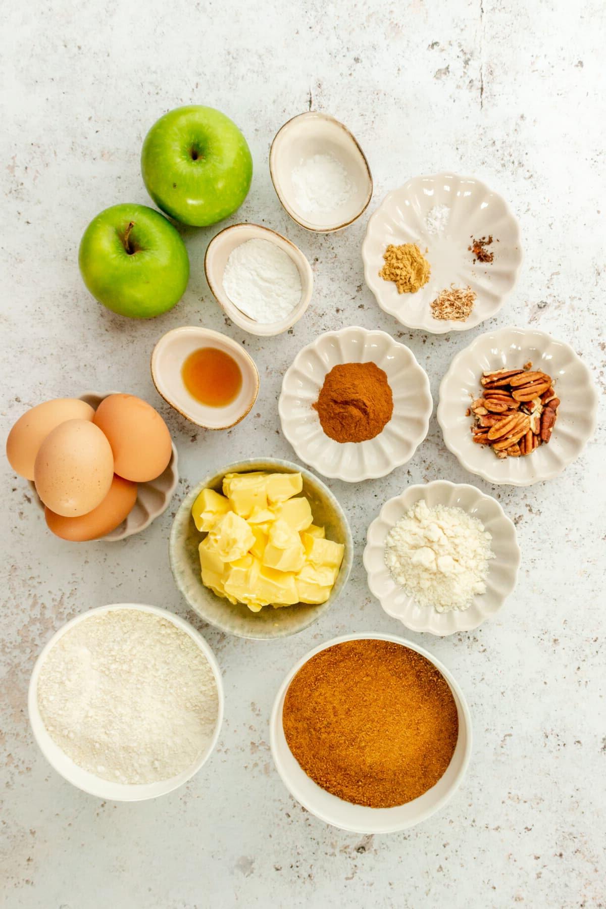 **Paläo Apfel-Zimt-Muffins** 3 Ingredients for paleo apple cinnamon muffins sit in a variety of bowls and plates on a light grey surface.