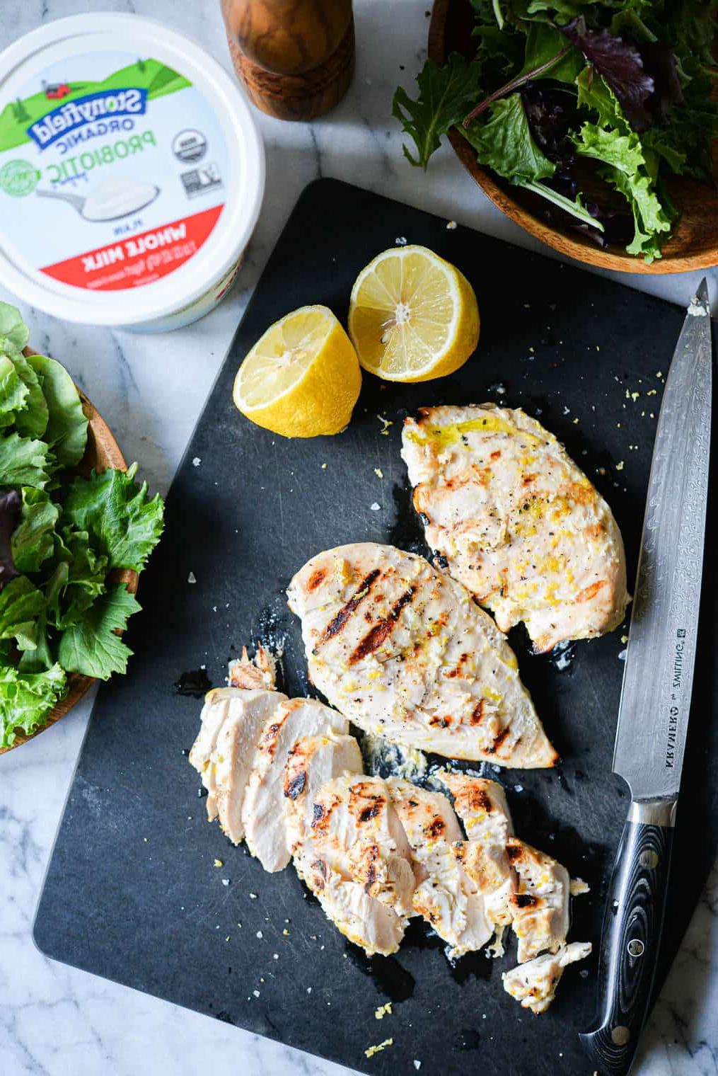 Zitronenpfeffer-Joghurt-Grillhähnchen 3 Drei Zitronen-Pfeffer-Joghurt-Hähnchenbrüste vom Grill auf einem schwarzen Schneidebrett neben einem Becher Naturjoghurt und einer halbierten Zitrone