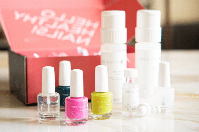 Dazzle Dry gegen Olive and June 2 Dazzle Dry Nagelset mit drei Nagellacken, einem Überlack und einem Entferner vor einer leuchtend rosafarbenen Box