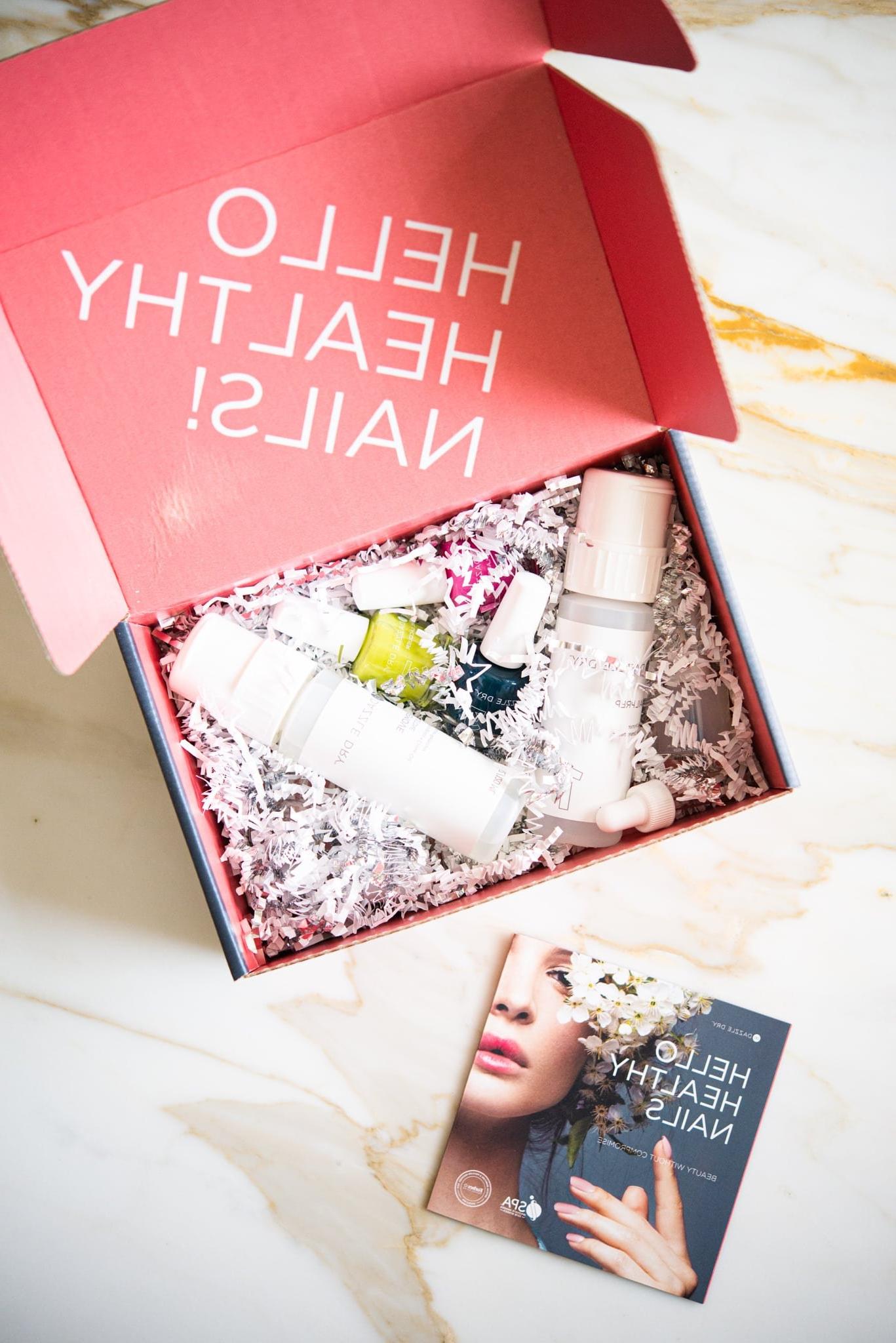Dazzle Dry Nail System Rezension 4 Eine rosa Box, gefüllt mit 3 verschiedenen Nagellackfarben, zwei klaren Lacken und einem Nagellackentferner, elegant platziert auf einer edlen Marmoroberfläche – das komplette Dazzle Dry System für zu Hause.