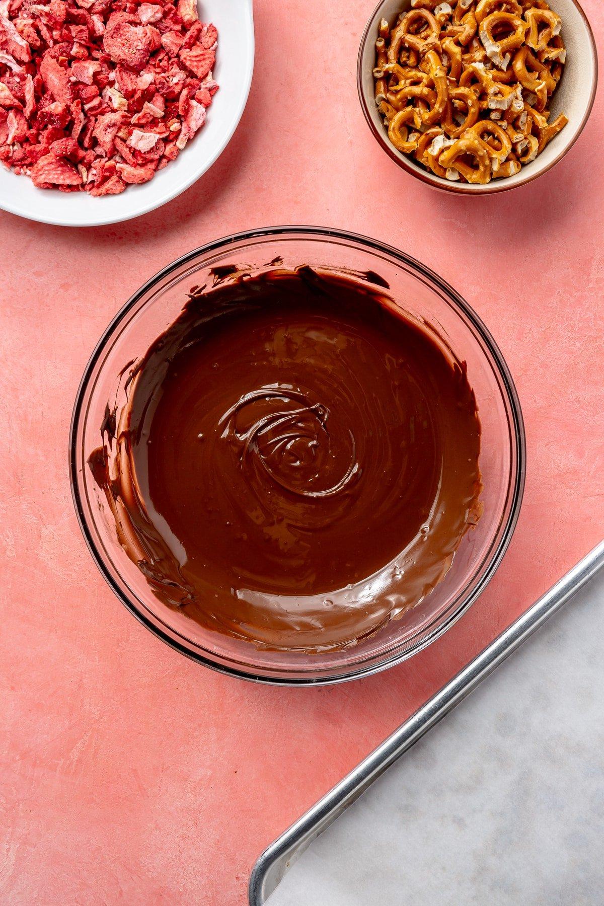 Geschmolzene Schokolade in einer Glasschüssel. Daneben stehen eine Schüssel mit Brezeln und eine Schüssel mit gefriergetrockneten Erdbeeren.