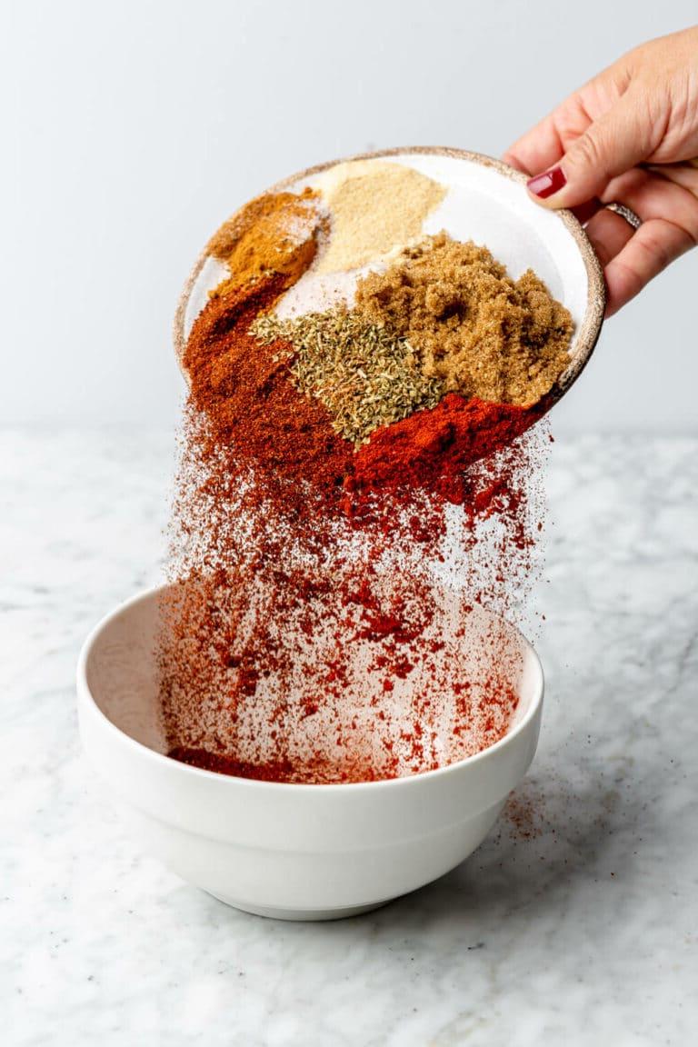 220928_Pork Tenderloin Rub-2 Eine Hand hält eine runde, flache Schale, aus der Gewürze in eine weiße Schüssel auf einer grau-weißen Marmoroberfläche gegossen werden.