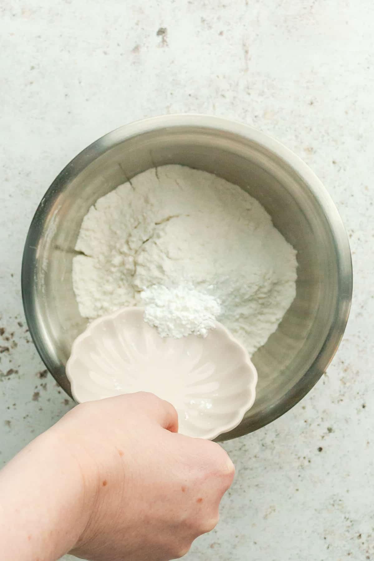 **Weiße Schokoladen-Pfefferminz-Kühlschrankkekse** 4 Natron wird über Mehl in einer Edelstahlschüssel auf hellem grauem Untergrund gegeben.
