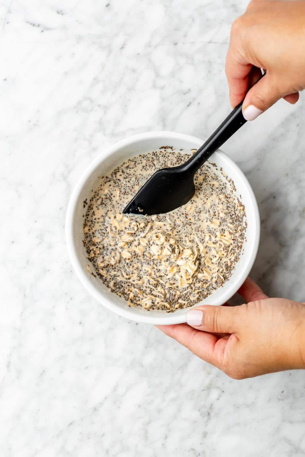 Schritt 1: Zutaten mischen Hand rührt mit einem kleinen schwarzen Spatel Haferflocken, Chia und Milch in einer Schüssel um.