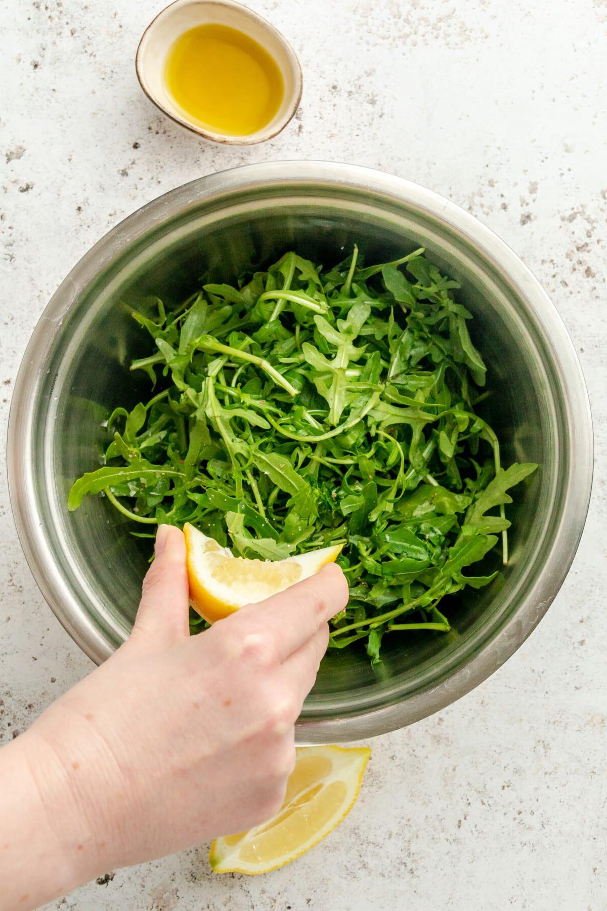 Rucola Salat mit Zitrone und Meersalz 4 Eine Zitronenspalte wird über einen Rucola Salat in einer Edelstahlschüssel auf hellem Hintergrund ausgepresst.