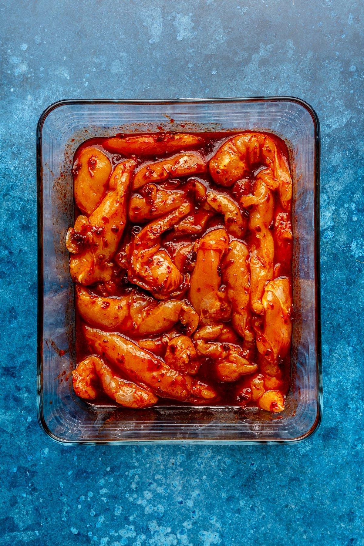 Harissa-Honig-Hähnchen-Bowls 4 Stücke von rohem Hähnchen, bedeckt mit der roten Flüssigkeit, liegen in einem rechteckigen Glasbehälter.