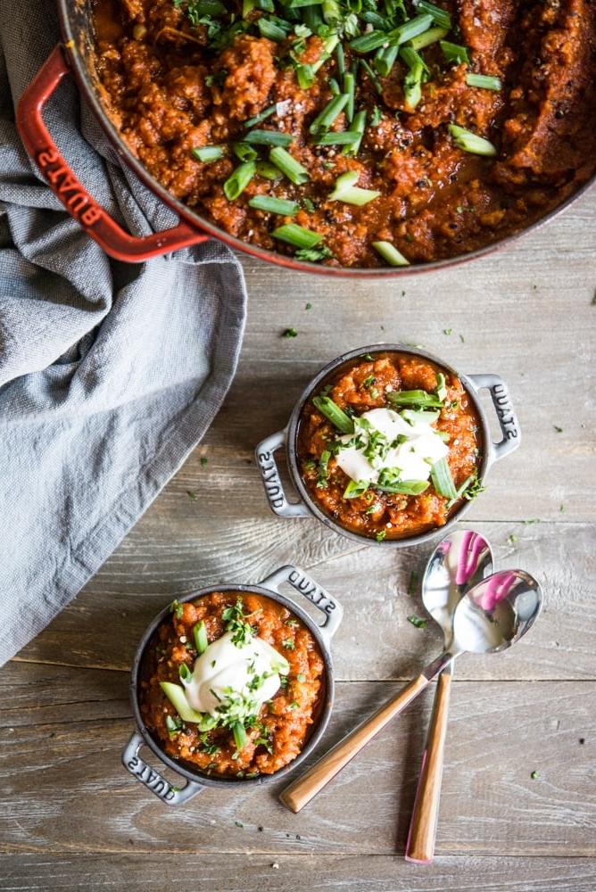 Ein großer roter Emailletopf gefüllt mit einem tief orangefarbenen Kürbis-Chili, garniert mit geschnittenen Frühlingszwiebeln, daneben zwei individuelle Portionen Chili, garniert mit einem Klecks Sauerrahm und geschnittenen Frühlingszwiebeln.
