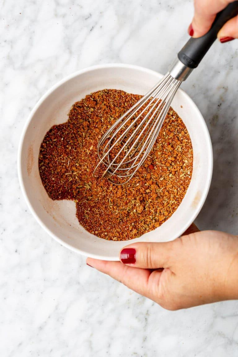 220928_Pork Tenderloin Rub-3 Eine Hand hält den Rand einer weißen Schüssel, die andere Hand hält einen Schneebesen mit schwarzem Griff. Eine Gewürzmischung wird in der weißen Schüssel verrührt.