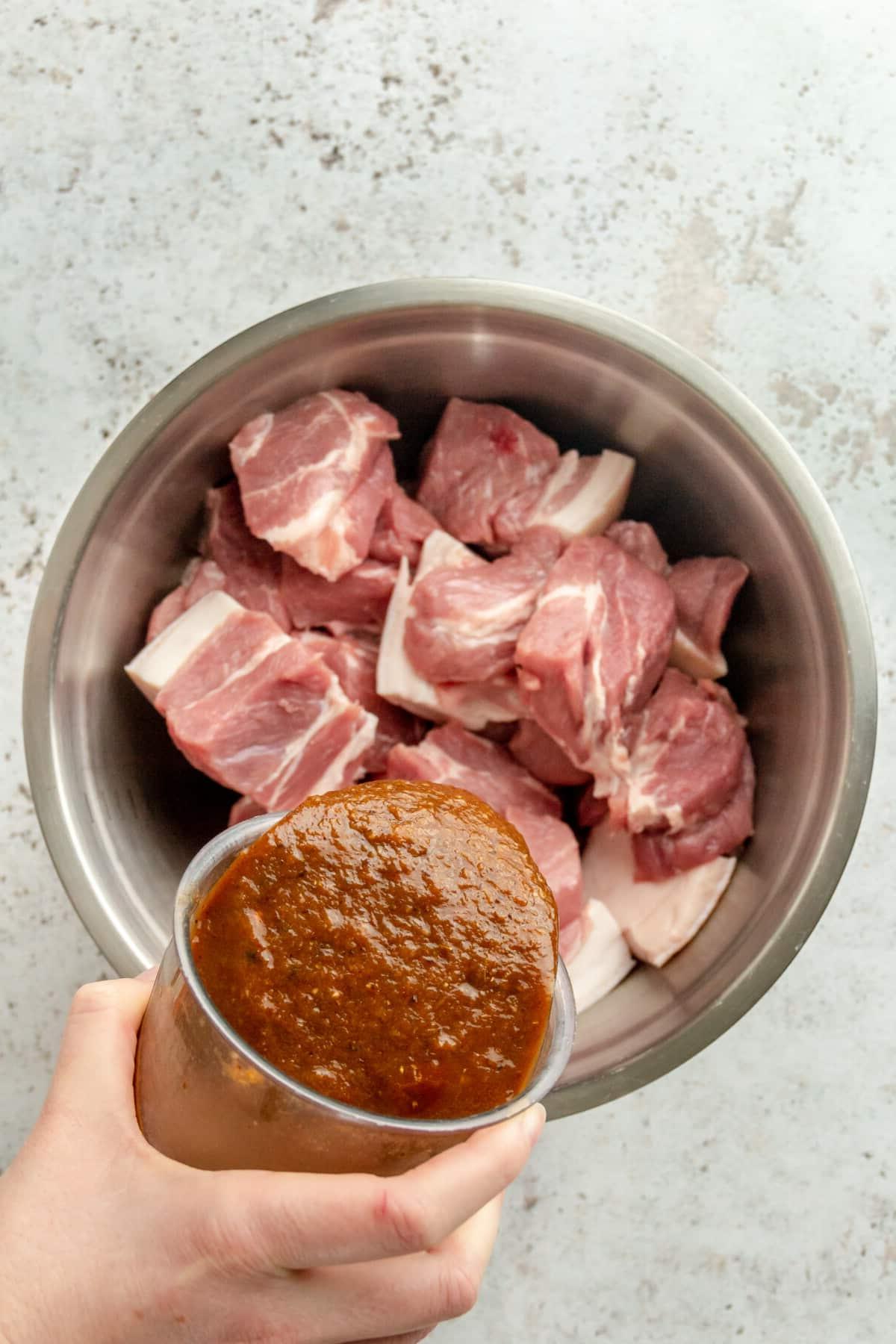 Tacos Al Pastor Rezept 5 Die vorbereitete Marinade wird über die Schweineschulterstücke in einer großen Edelstahlschüssel auf hellem Hintergrund gegossen.