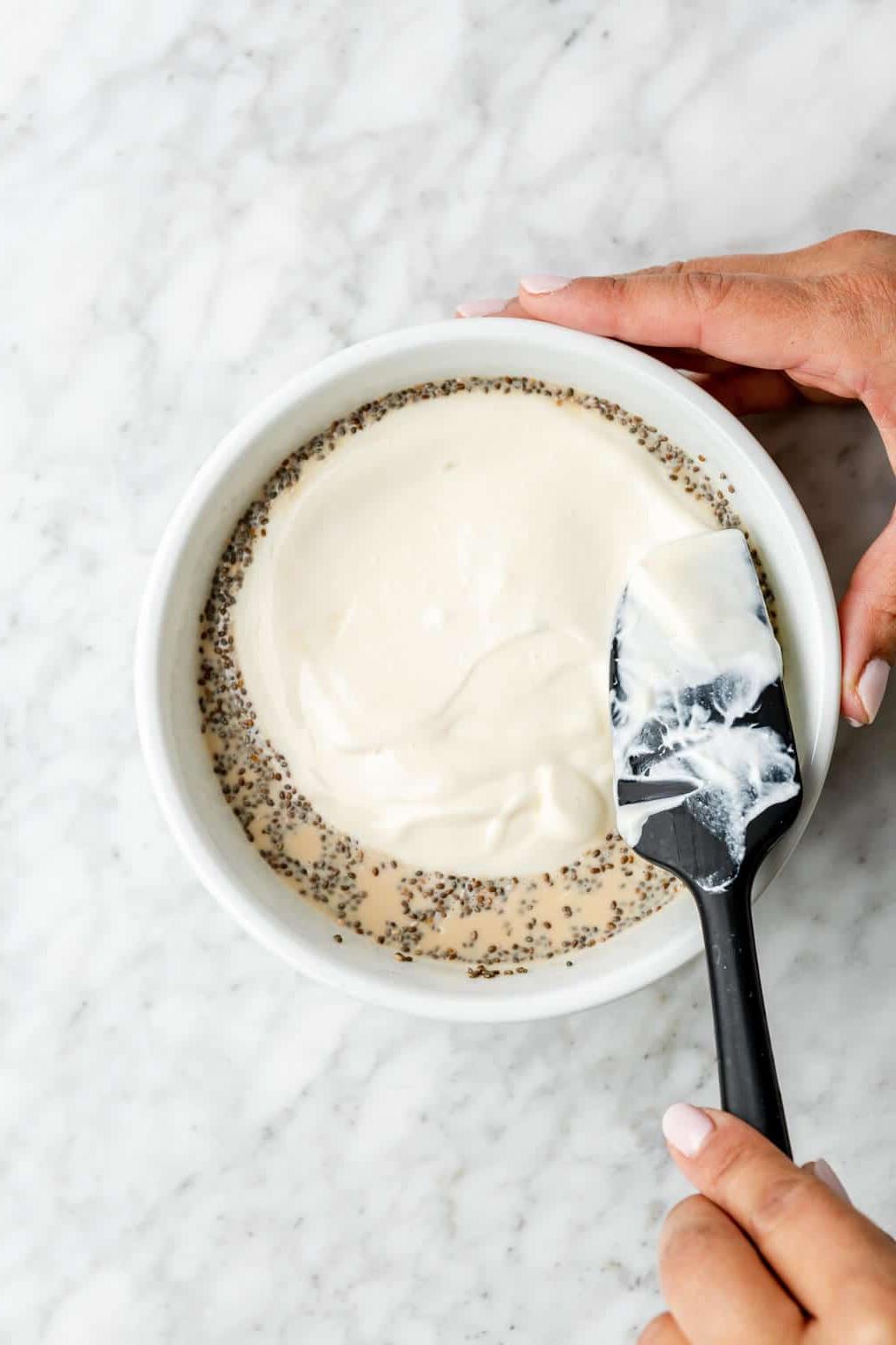 Schritt 3: Joghurt-Schicht hinzufügen Hand streicht mit einem schwarzen Spatel Joghurt über die Haferflockenmischung in einer Schüssel.
