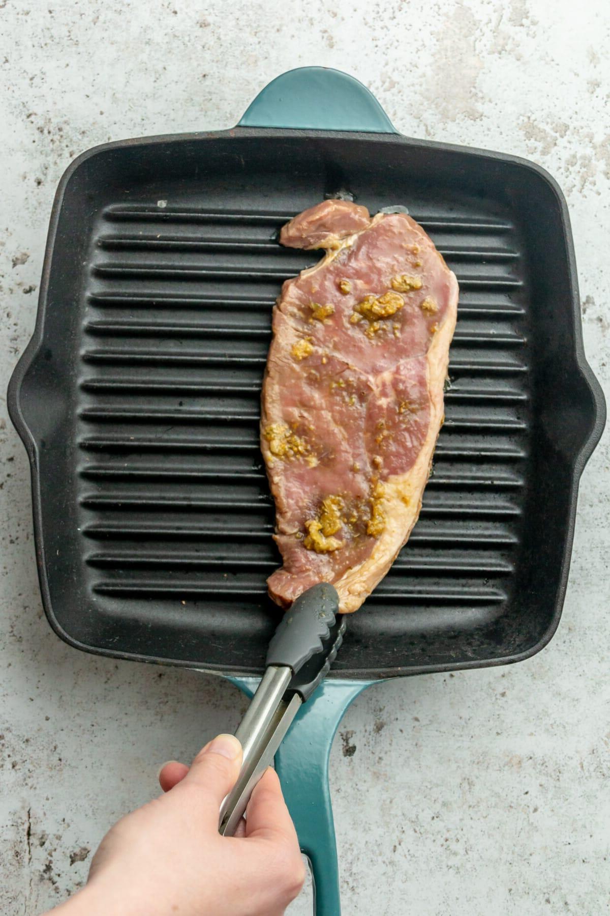 Carne Asada Tacos Rezept mit Adobo-Limetten-Marinade 6 Ein Stück Steak wird auf eine heiße Grillpfanne auf einer hellgrauen Oberfläche gelegt.