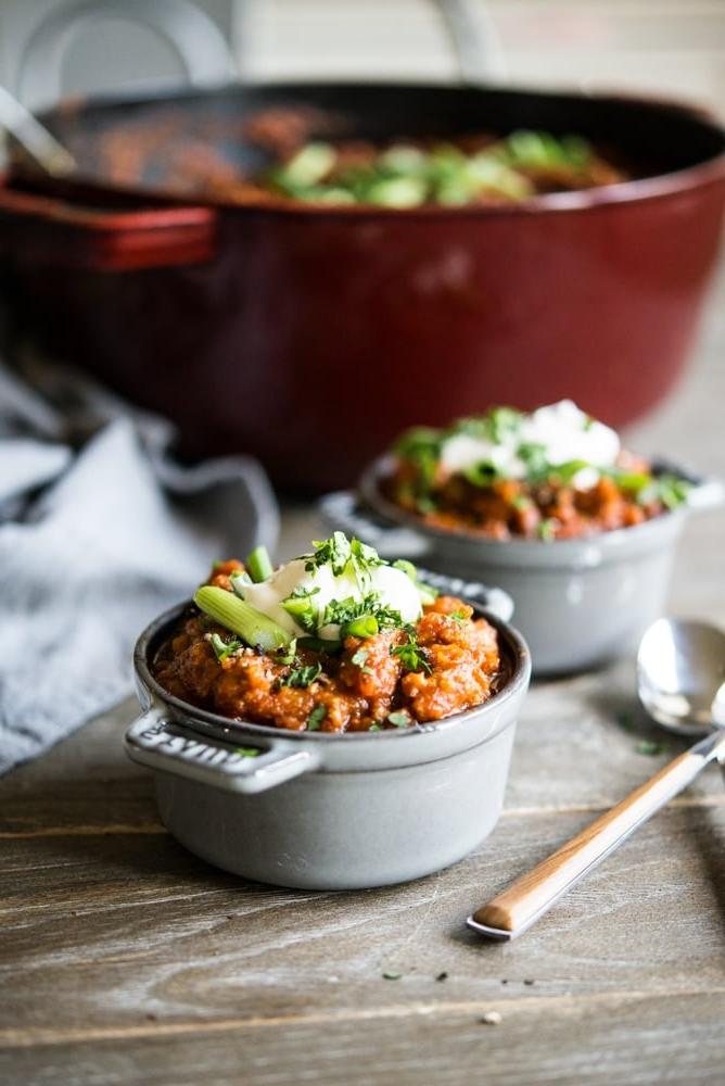 Ein großer roter Emailletopf gefüllt mit einem tief orangefarbenen Kürbis-Chili, garniert mit geschnittenen Frühlingszwiebeln, daneben zwei individuelle Portionen Chili, garniert mit einem Klecks Sauerrahm und geschnittenen Frühlingszwiebeln.
