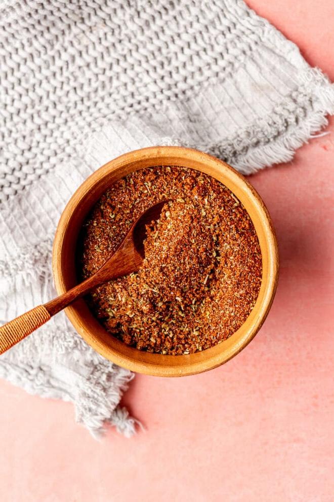 Beste Trockenmarinade für Schweinefilet 4 Draufsicht auf eine braune Tonschale mit einem kleinen Holzlöffel, gefüllt mit einer Gewürzmischung. Die Schale steht auf einer rosa Oberfläche mit einem hellgrauen, strukturierten Leinen.
