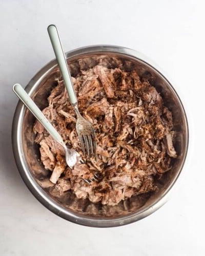 Eine Schüssel mit zerkleinertem Schweinefleisch, bestreut mit Gewürzen, und zwei Gabeln in der Schüssel zum Mischen