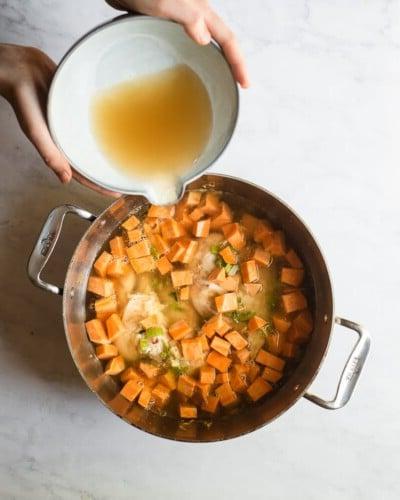 210423_Healing Tumeric Soup-23 Eine Frau gießt Brühe in einen Edelstahltopf mit Hähnchen, Sellerie, gewürfelten Süßkartoffeln und Gewürzen