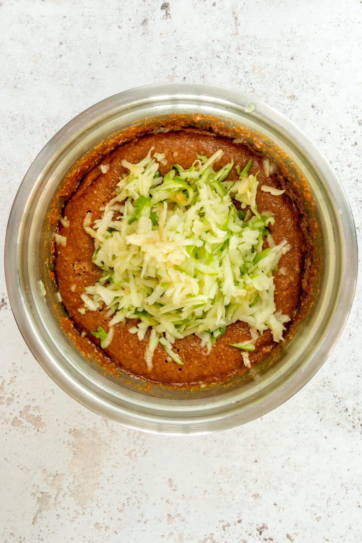 **Paläo Apfel-Zimt-Muffins** 6 Grated apple sits on top of a paleo cinnamon muffin batter in a stainless steel bowl on a light grey surface.