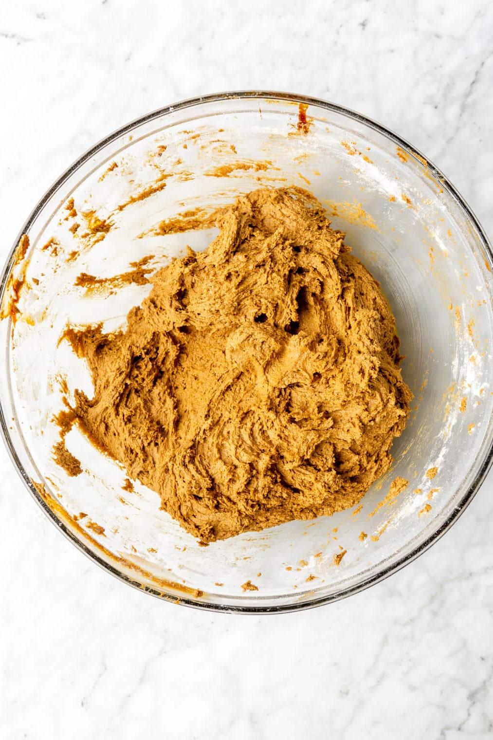 220817_Rum Raisin Cookies-28 Gemischter Teig in einer Glasschüssel auf einer grau-weißen Marmoroberfläche. Der Teig ist goldbraun.