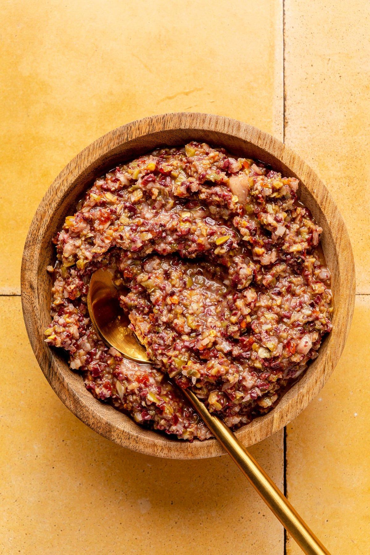 Oliven-Tapenade 6 Oliven-Tapenade in einer hölzernen Servierschale. Ein goldfarbener Löffel liegt daneben.
