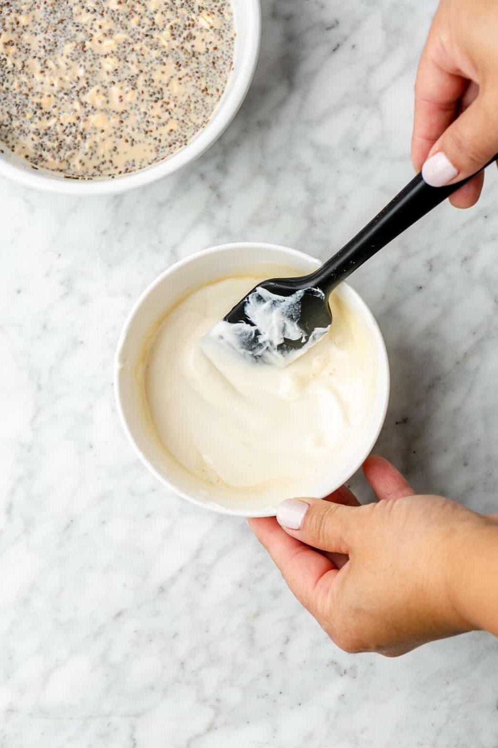 Schritt 2: Joghurt-Mischung zubereiten Hand rührt mit einem schwarzen Spatel Joghurt und Ahornsirup in einer kleinen weißen Schüssel zusammen.
